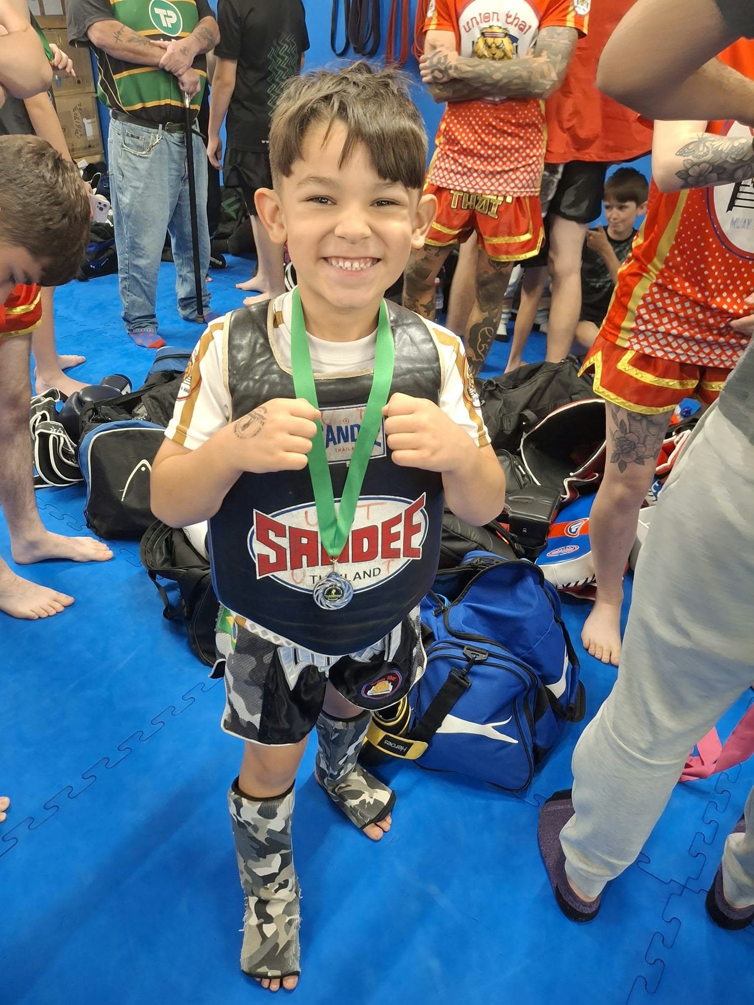 Young Union Thai fighter smiling with a medal and a Sandee chest guard at a kids event