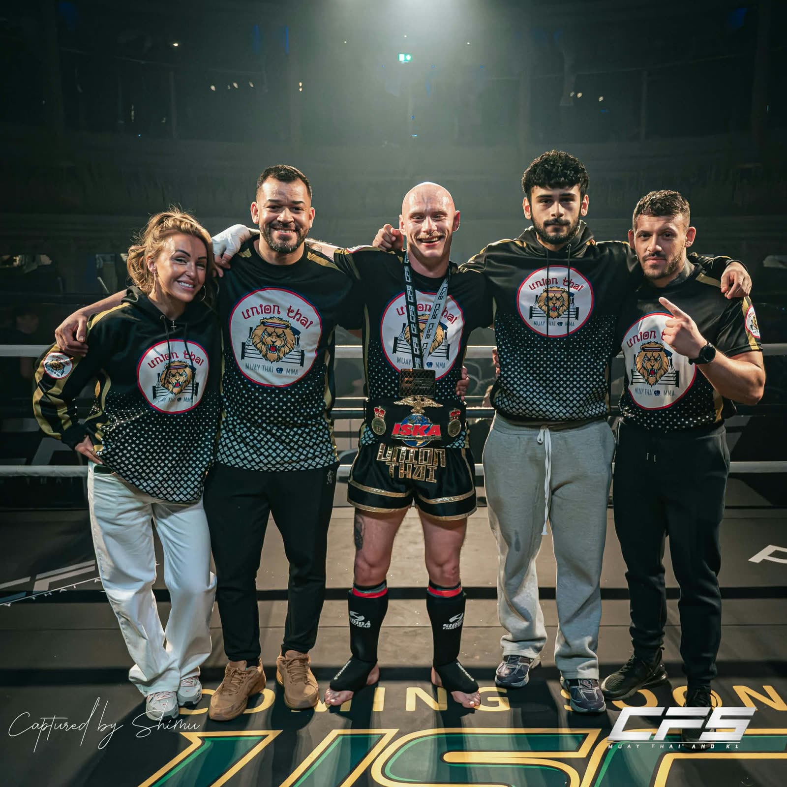 Five Union Thai coaches and corner team posing in the ring with a fighter wearing a championship belt